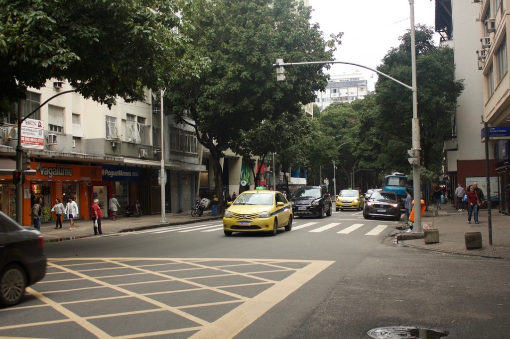 A street scene featuring cars, including yellow taxis, with pedestrians walking on the sidewalk amidst urban buildings and trees.