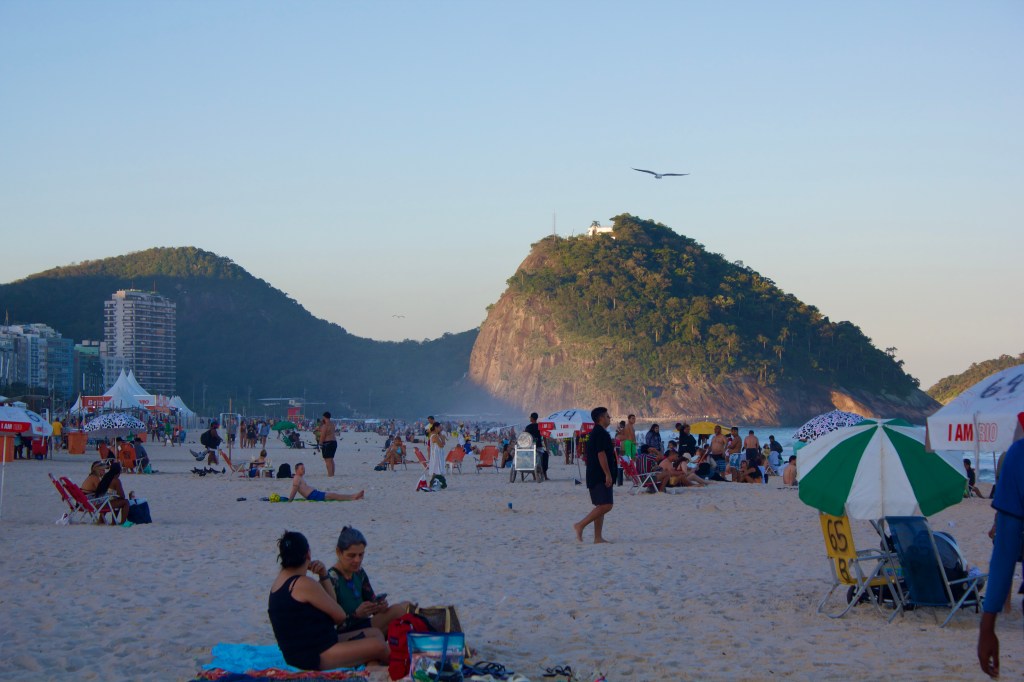 A crowded beach scene with people lounging on the sand, colorful umbrellas, and a tall hill in the background, under a clear sky.
